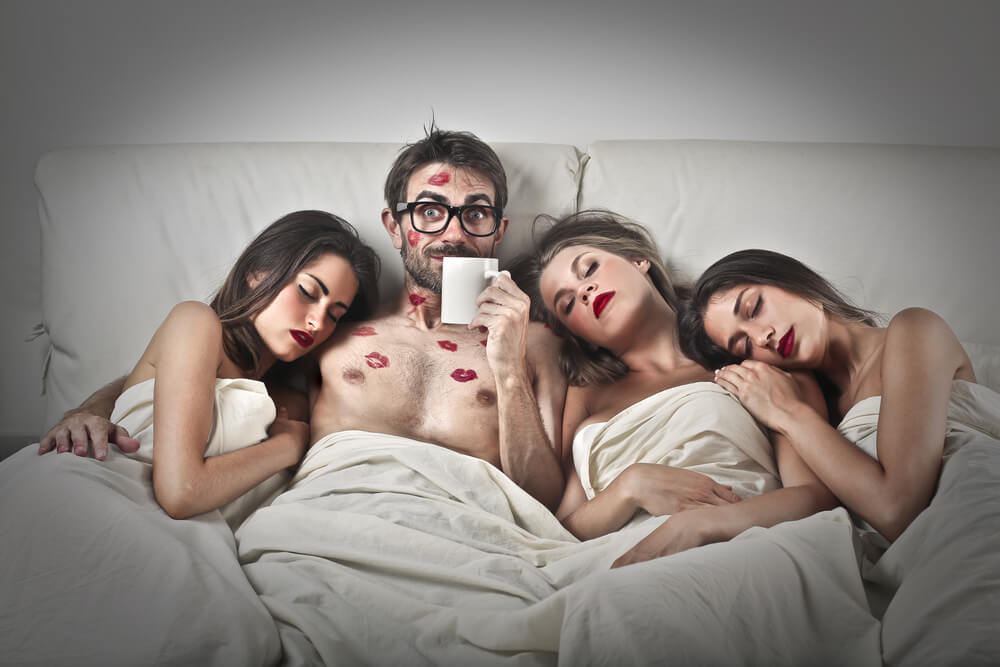 A guy with three beautiful women lying in bed. The man has kissing marks on his face and chest.