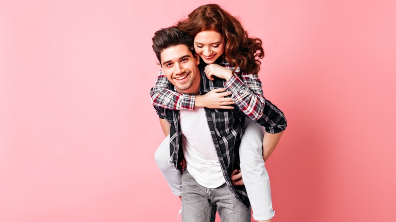 asexual guy happy to give his pretty ginger girlfriend a piggyback ride in front of a pink background